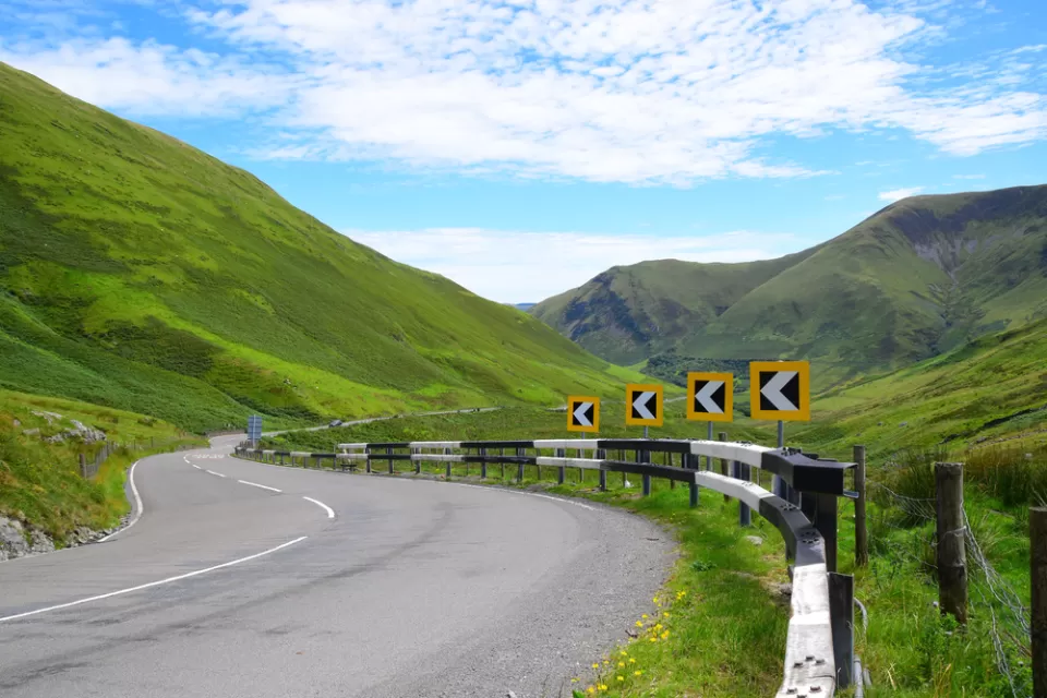 Asphalt road with yellow and black turn warning signs winding through green Welsh mountain landscape